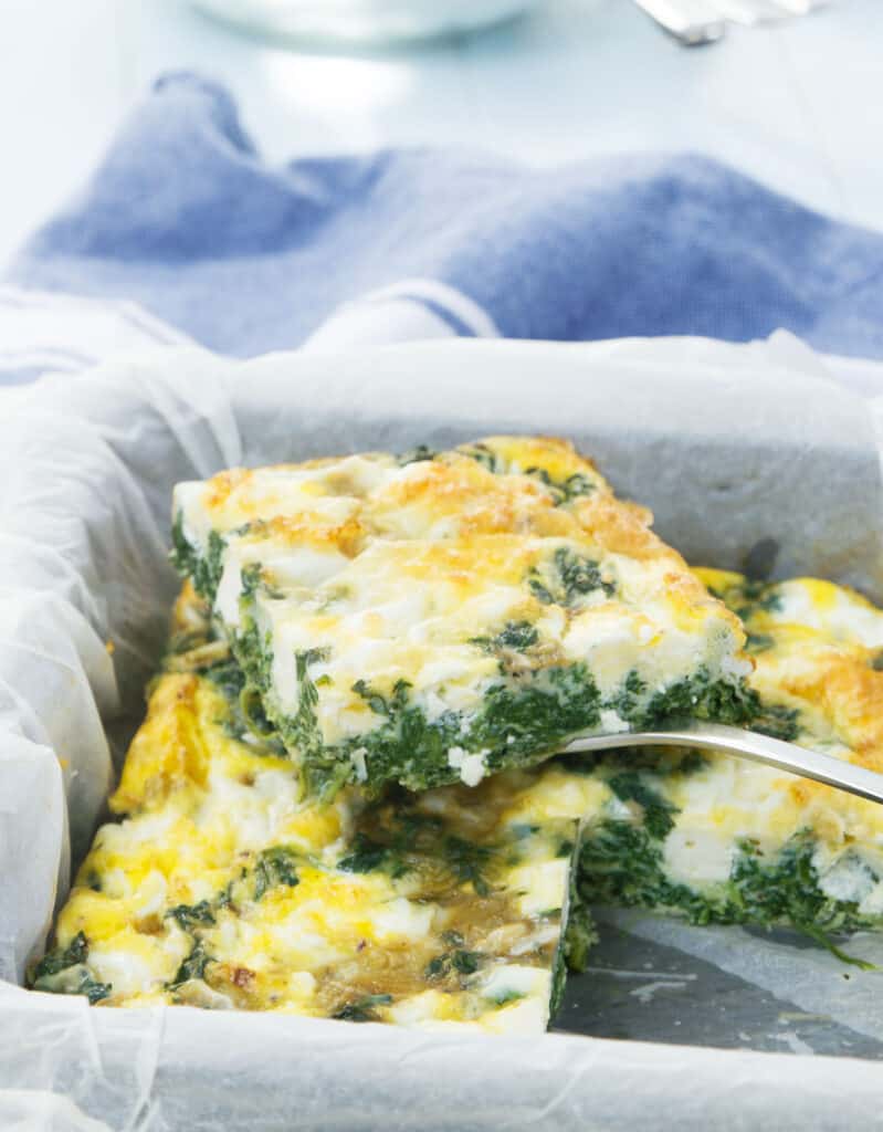 Close-up of a spinach frittata in the squared pan over a light blue background.