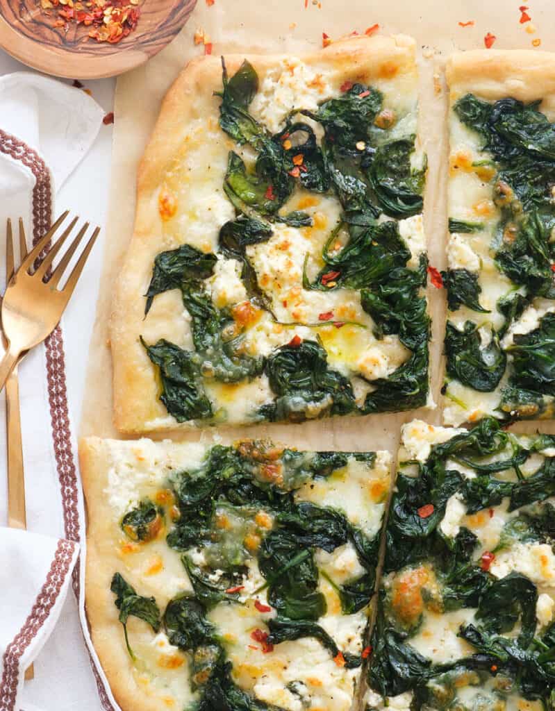 Top view of a large spinach pizza cut into slices and scattered with red chili flakes.