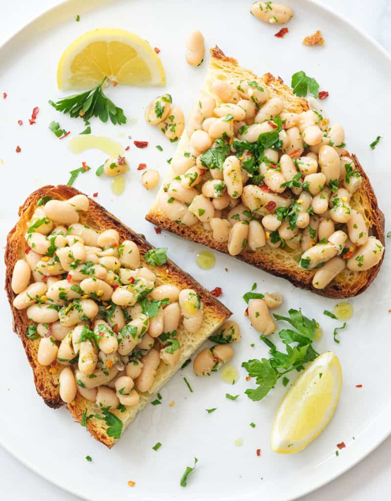 Top view of bread and beans served on a large white plate with lemon wedges and fresh parsley.