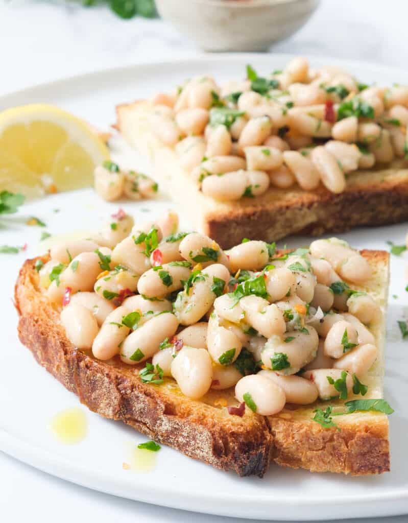 Close-up of a two slices of bread with beans, parsley and lemon over a white plate.