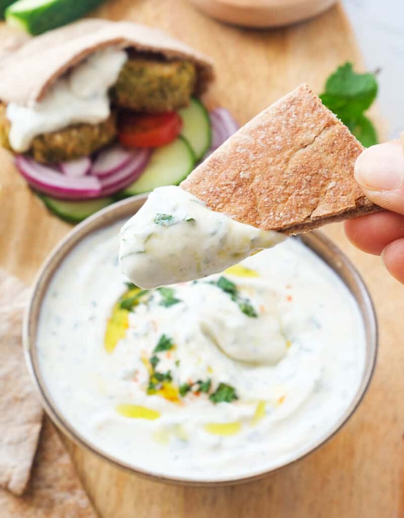 Close-up of a piece of pita bread dipped into the mint yogurt sauce. pita bread with falafels in the background.