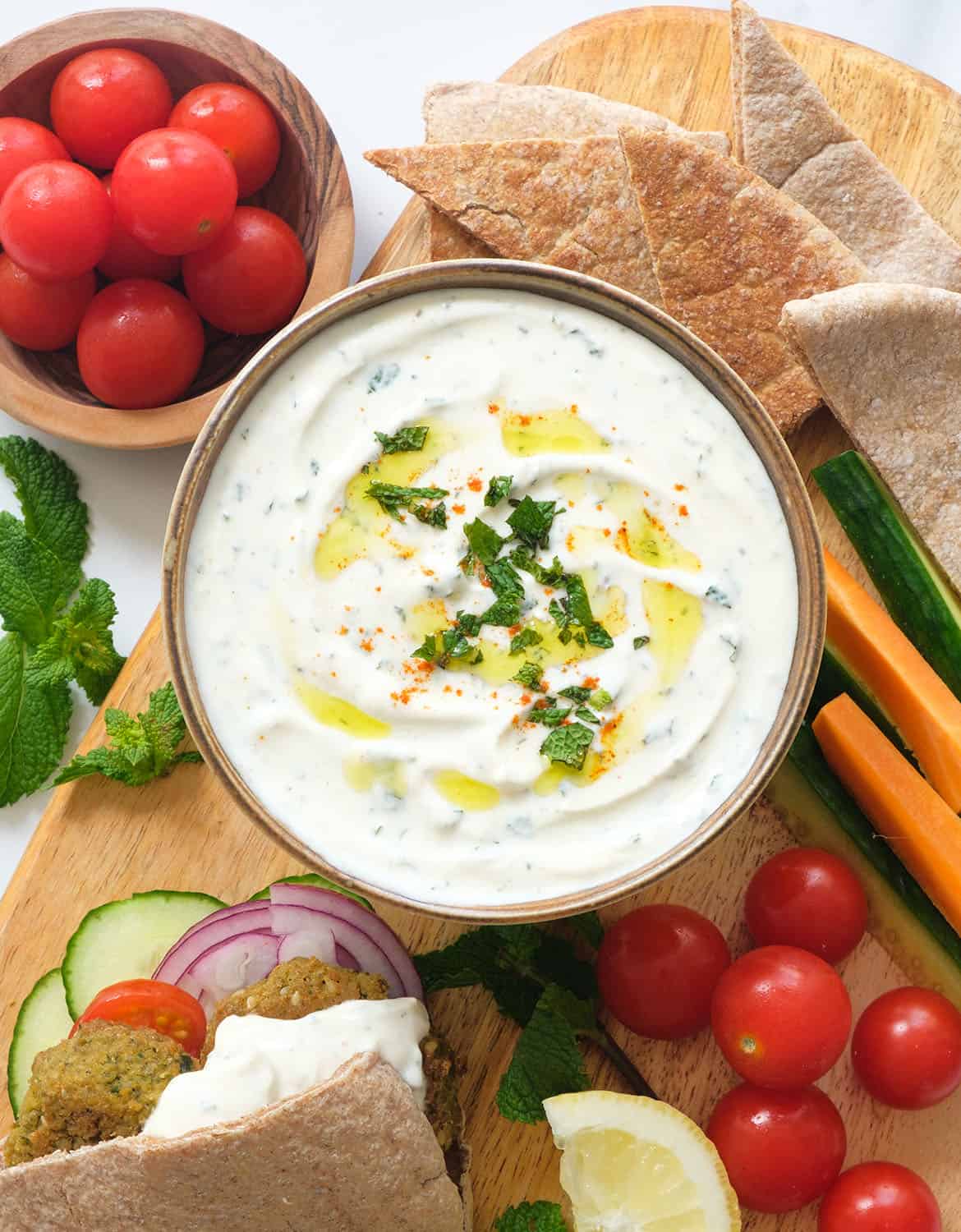 Top view of a bowl full of creamy mint yogurt sauce served with falafels, pita bread, cherry tomatoes and veggie sticks.