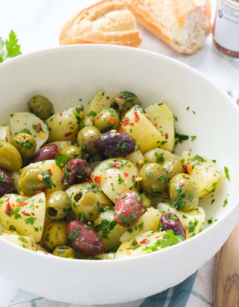 Close-up of a white bowl full of yummy salad with olives, potatoes, herbs and chili flakes.