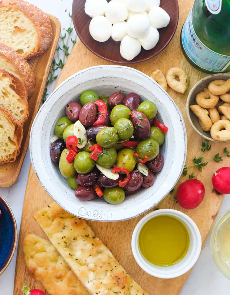 Top view of a serving suggestion showing a bowl full of spicy olives on a wooden platter full of other Italian appetizers and finger food.