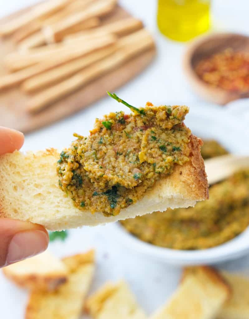 Close-up of a crusty piece of bread with green olive tapenade. Breadsticks and bread in the background.