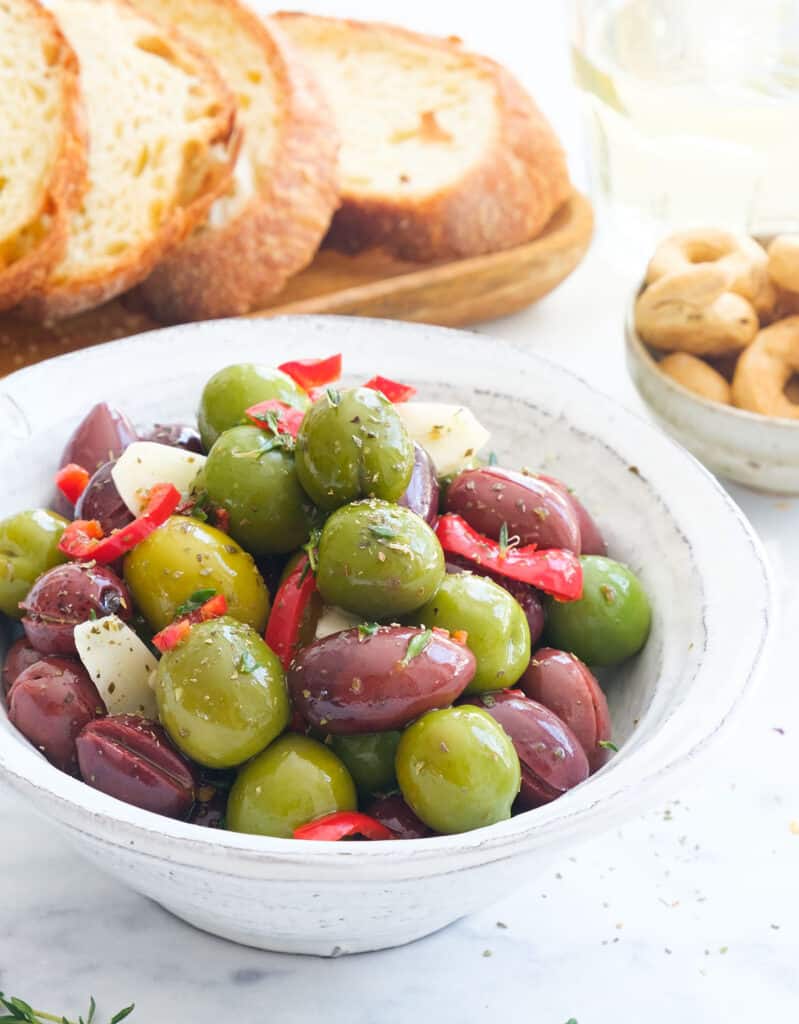 Close-up of a white bowl full of mixed Italian spicy olives served with bread and taralli. 