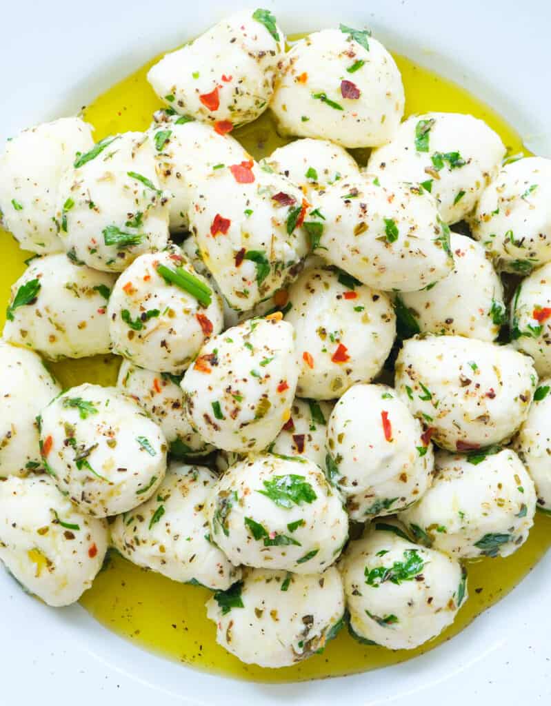 Close-up of plenty of baby mozzarella balls in olive oil with herbs over a white plate.