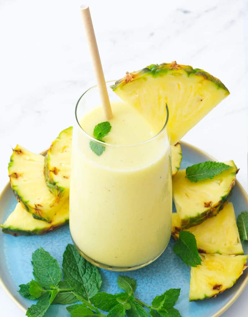 A glass full of creamy pineapple banana smoothie served with pineapple slices and mint leaves.