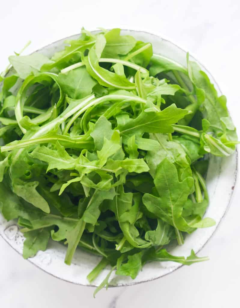 Top view of a white bowl full of fresh baby arugula star ingredients in this collection of arugula recipes.