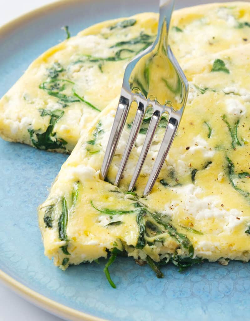 Close-up of a fork pinch a slice of soft arugula frittata with feta over a light blue plate.