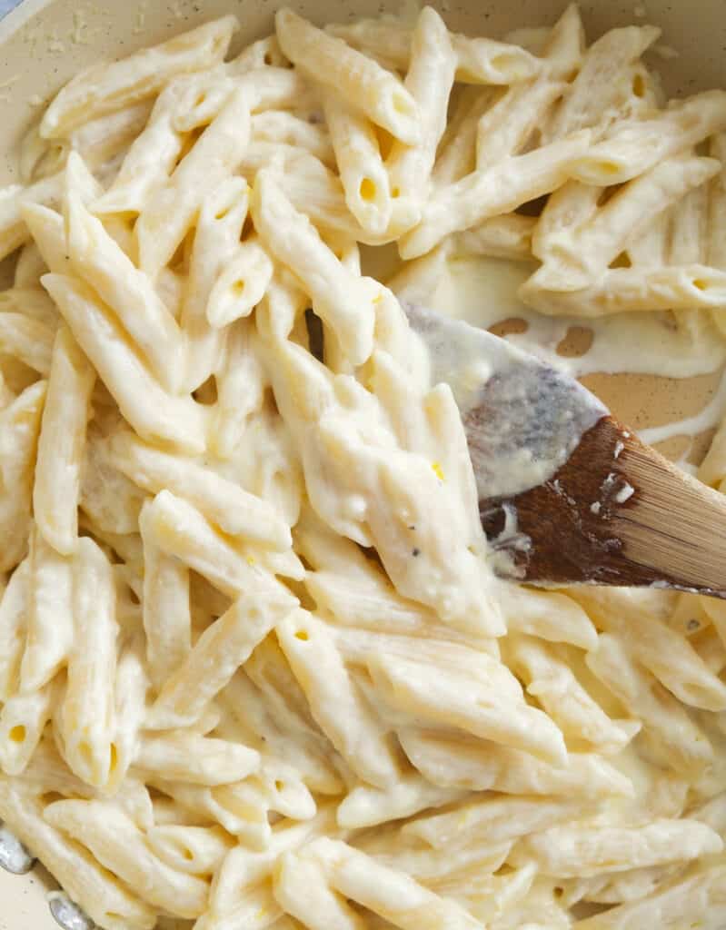 Top view of a wooden spoon stirring the creamy penne pasta in a skillet.