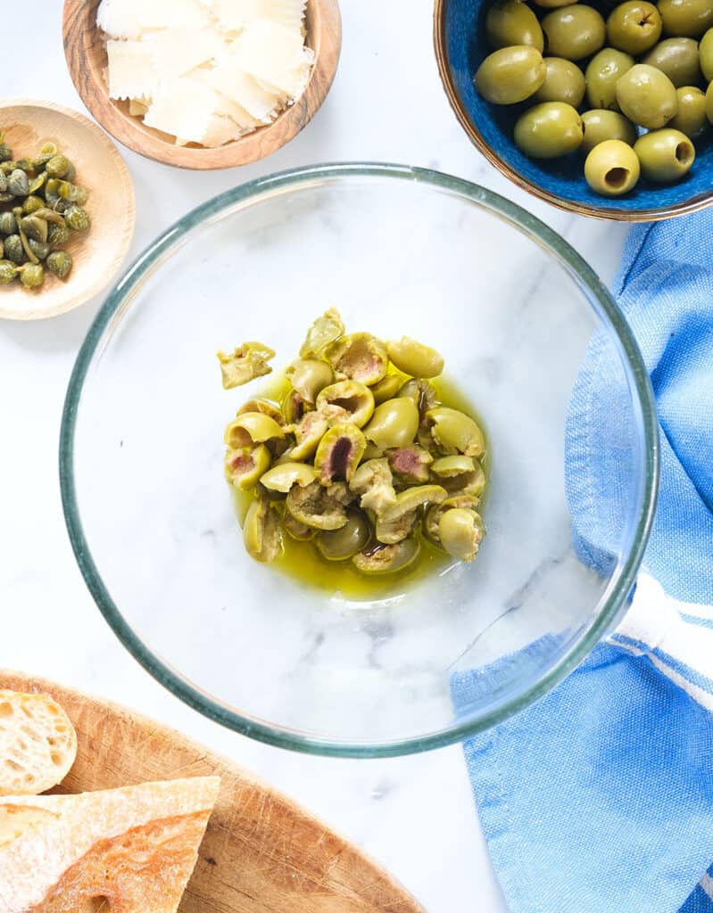 Top view of a glass bowl containing torn green olives and olive oil.