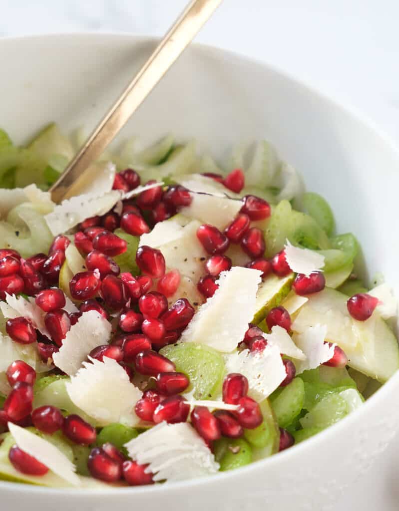 Close-up of a white salt bowl full of celery salad garnished with red juicy arils.
