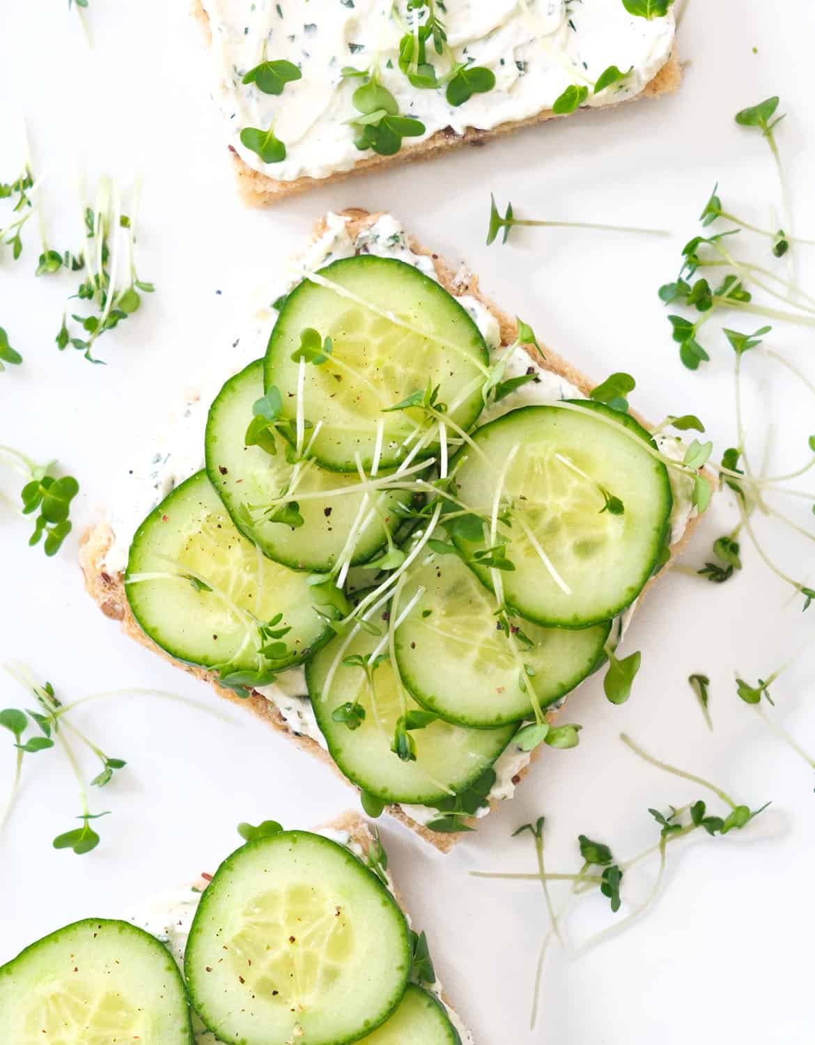 Top view of a few squared cream cheese sandwiches topped with sliced cucumber and English cress.