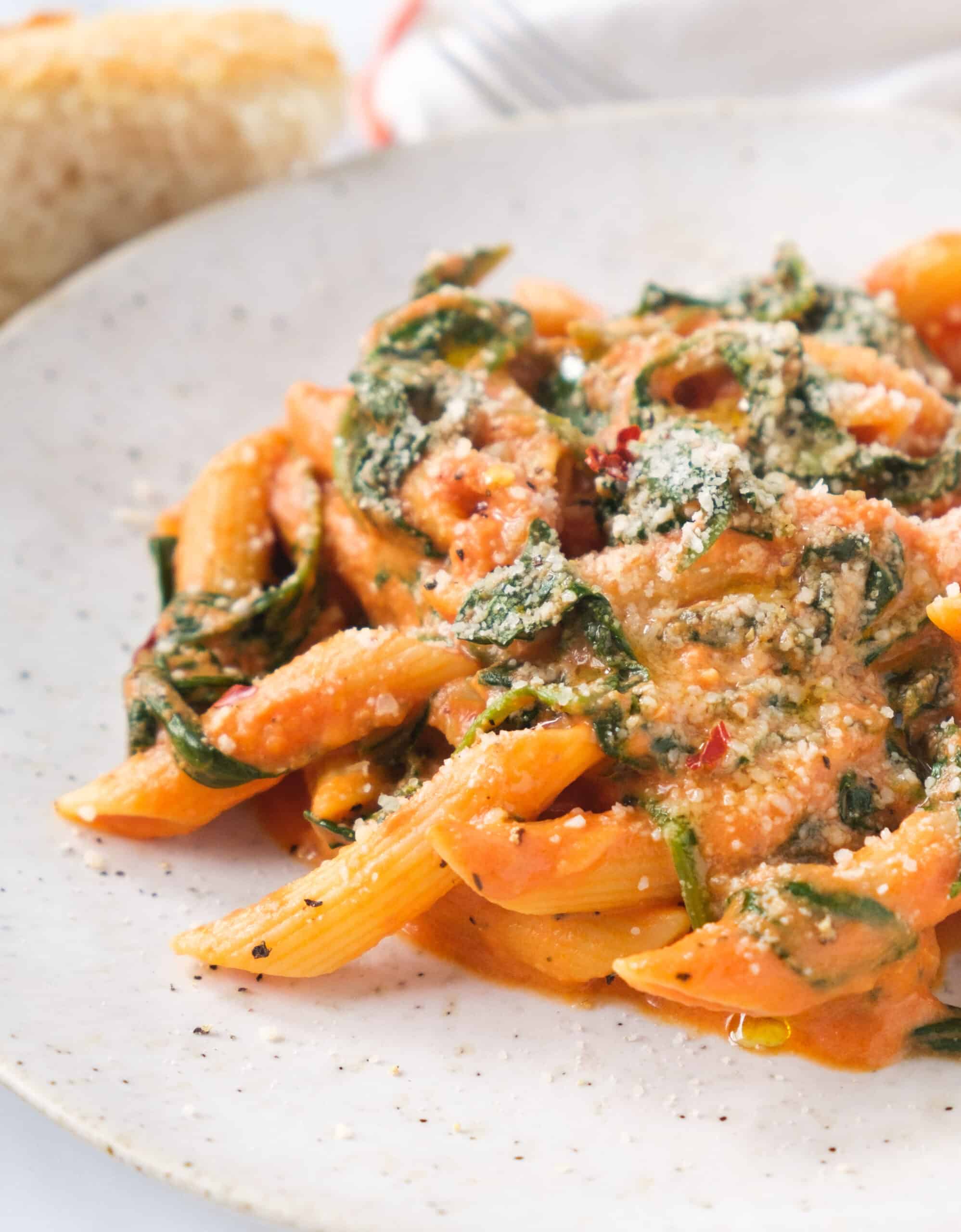 Close-up of a white plate with a serving of pasta with creamy tomato spinach sauce served with grated parmesan cheese.
