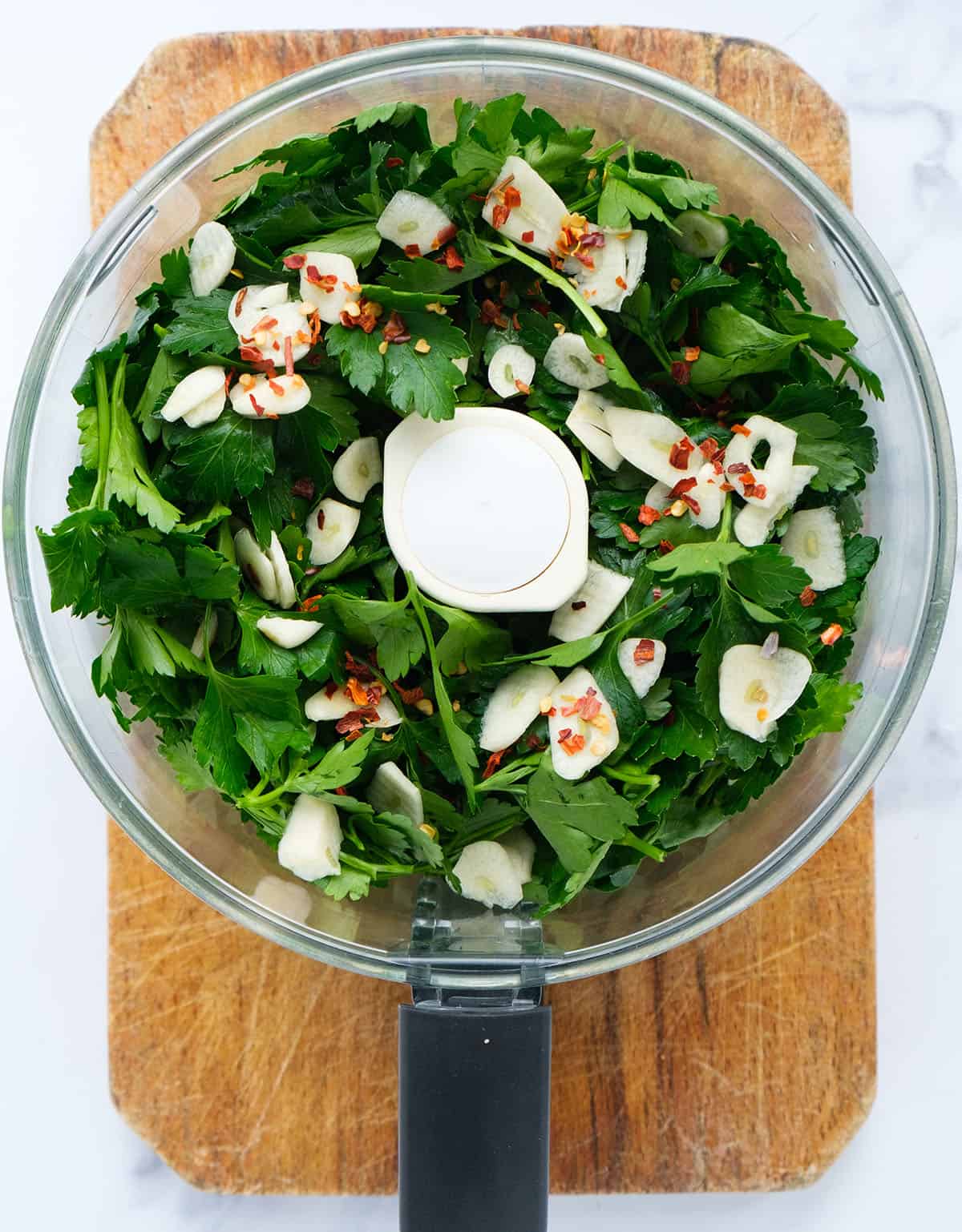 Top view of a food processor full or parsley leaves, garlic and chili flakes.