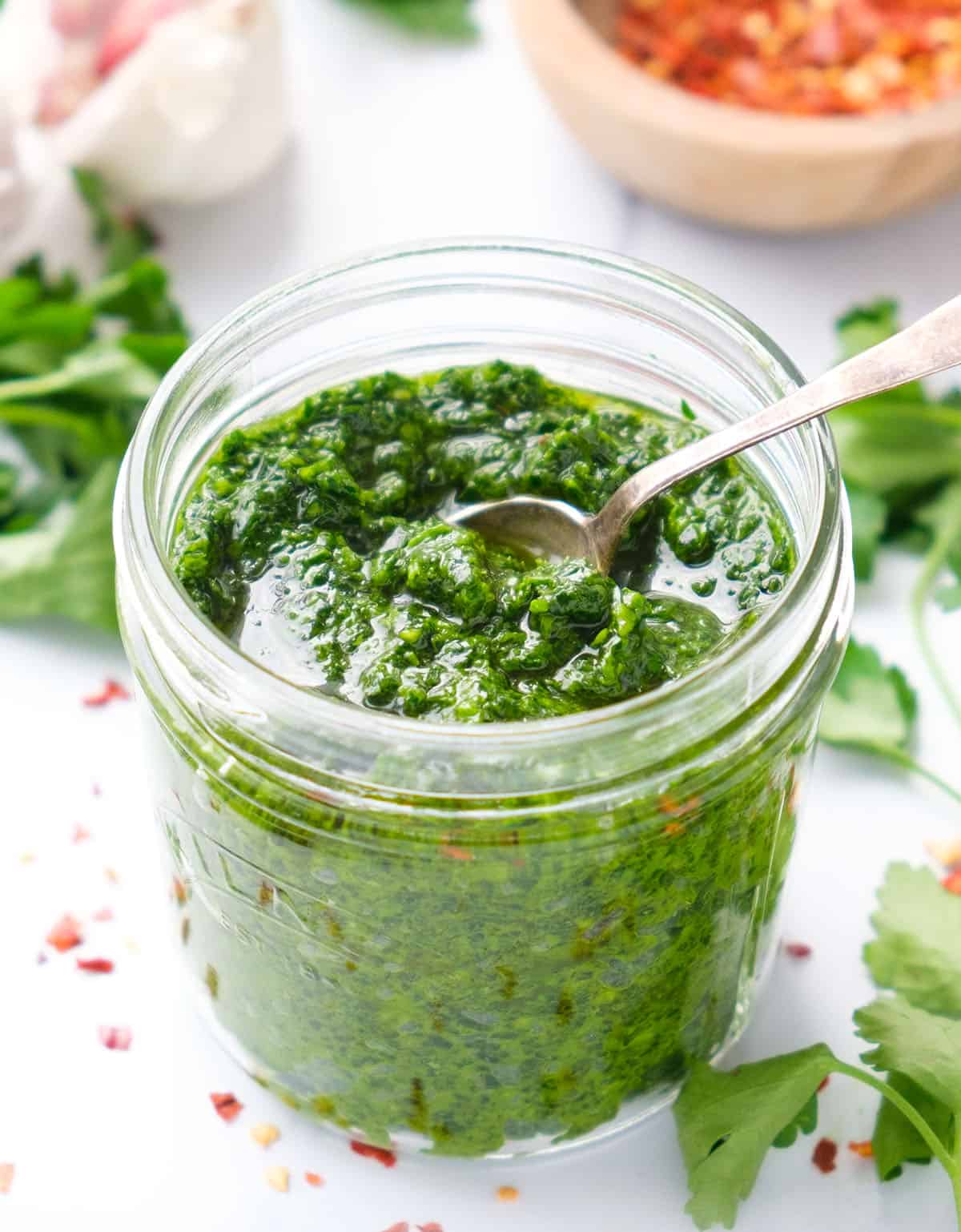 Close-up of a glass jar full of parsley pesto, parsley leaves and some chili flakes in the background.
