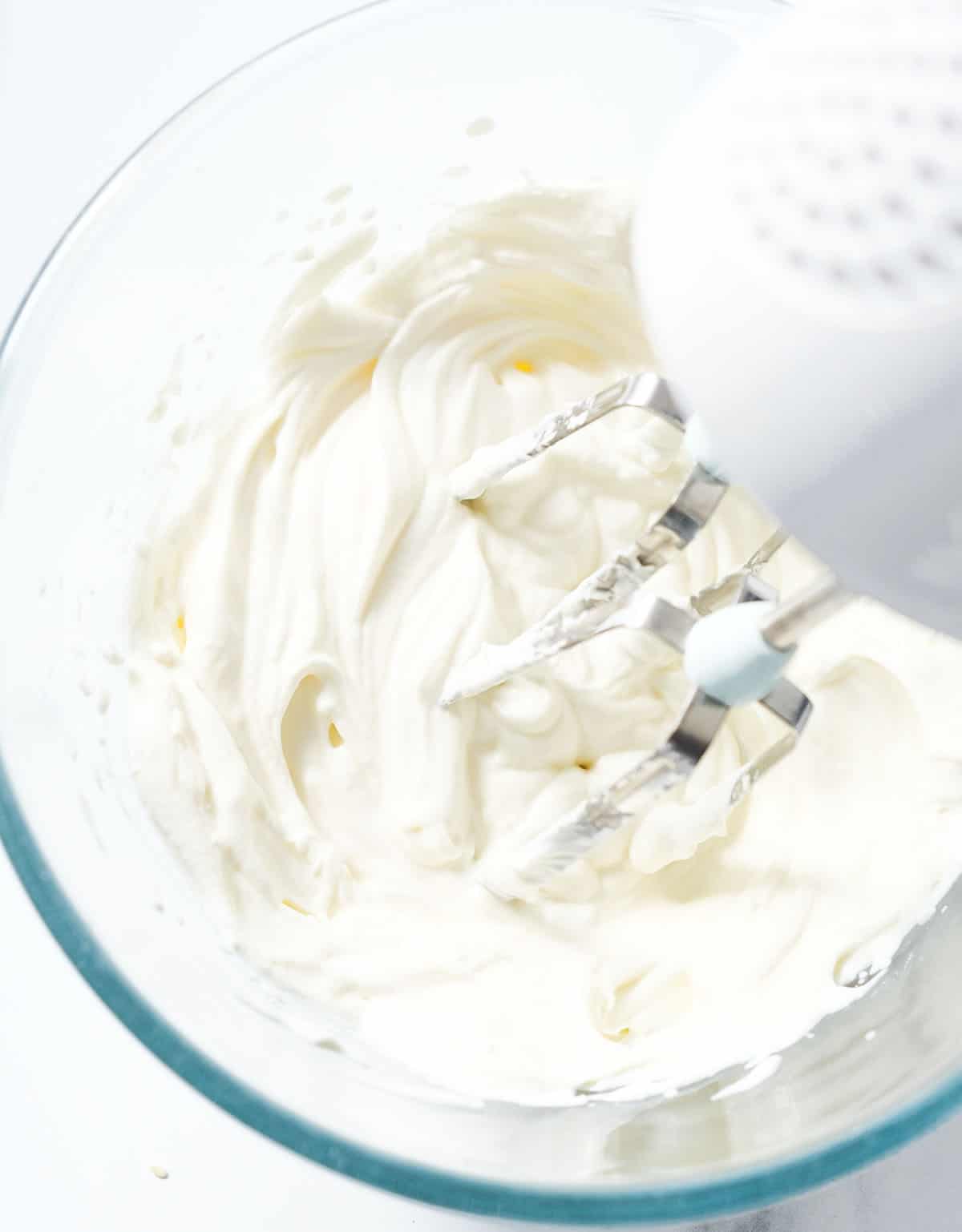 An electric hand beater beating cream cheese in a glass bowl over a white background.