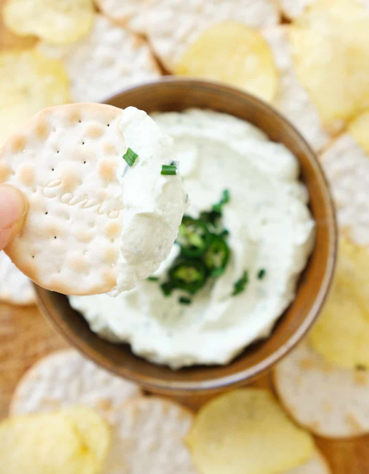 Close-up of a round cracker with jalapeño cream cheese and more dip, chips and crackers in the background. 