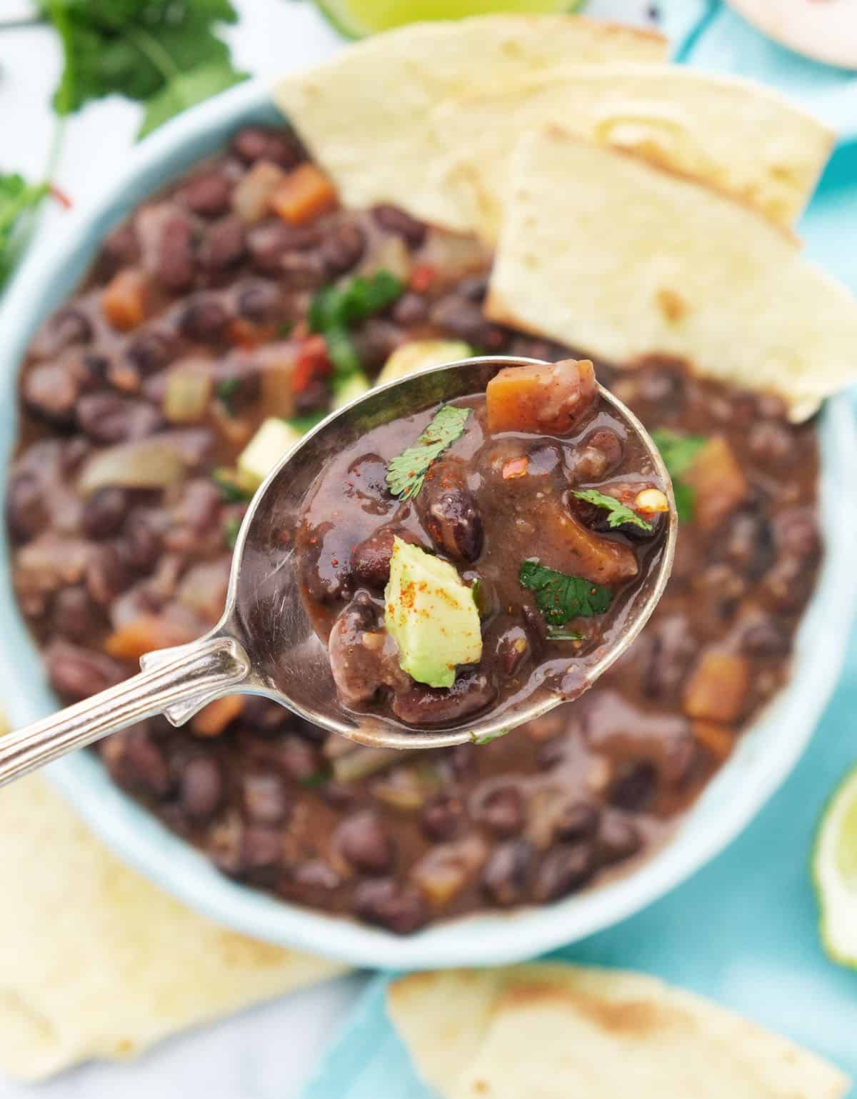 Close-up of a spoon full of vegan black bean soup and a bowl full of soup in the background.