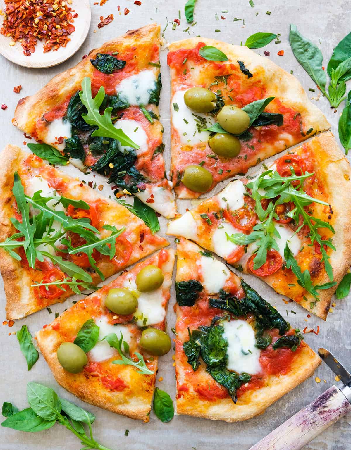 Top view of a large round pizza cut into slices with different pizza toppings like olives, rocket, and  spinach.