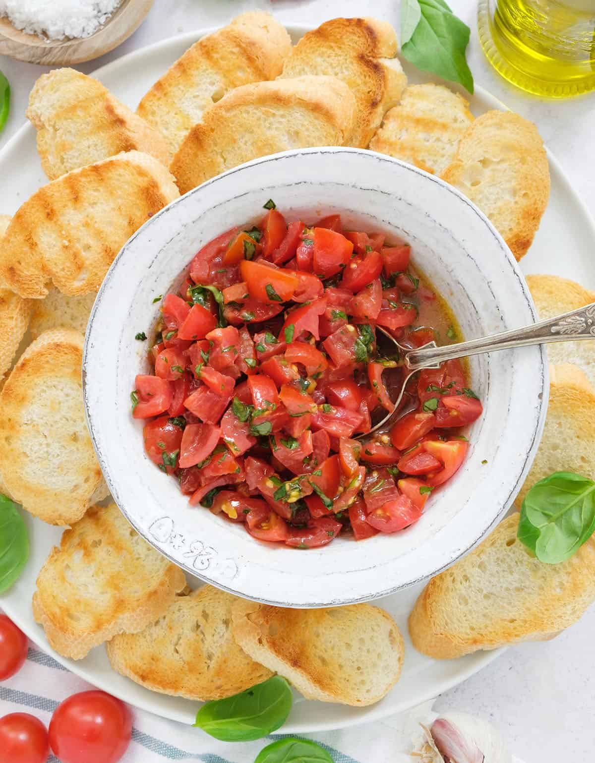 Top view of a white bowl full of tomato bruschetta dip with basil served with grilled slices of baguette.