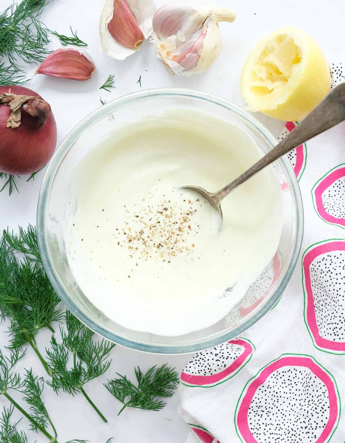 Top view of a glass bowl full of yogurt dressing. Lemon, garlic cloves and dill in the background.