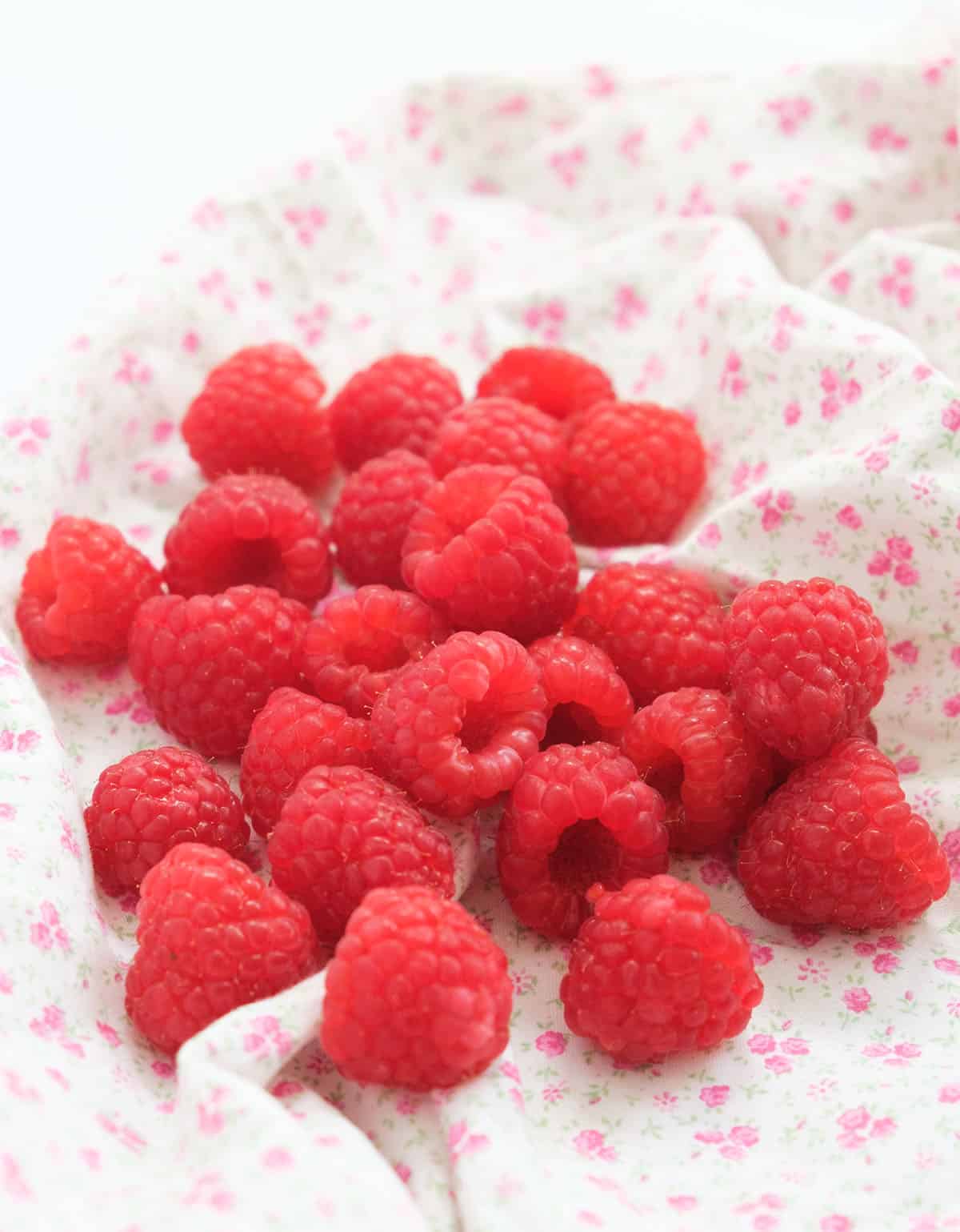 Close-up of a handful of fresh raspberries on a white tea towel decorated with small pink flowers.