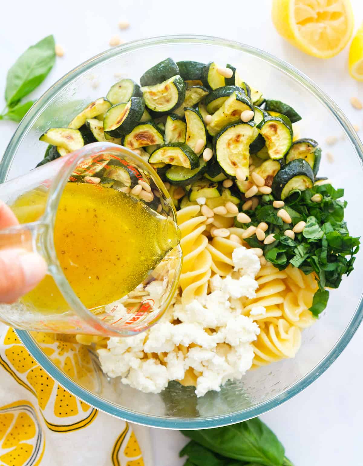 Top view of a glass jug pouring dressing over a bowl full of zucchini pasta salad.