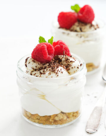 Close-up of two small glass jars full of cream cheese mousse and garnished with raspberries over a white background.
