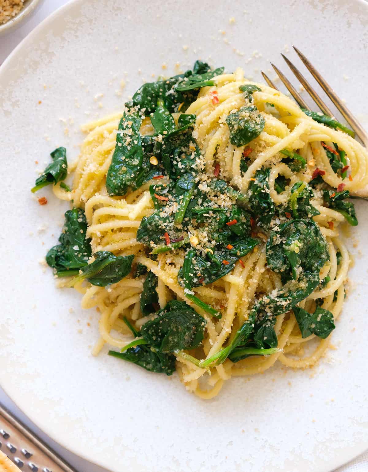 Top view of a white plate full of spinach spaghetti with garlic breadcrumbs and chilli flakes.