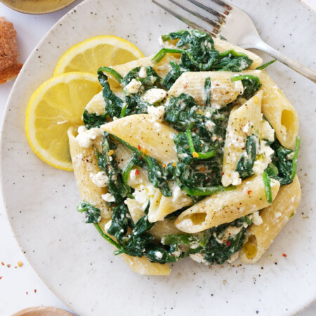 Top view of a white plate full of spinach feta pasta served with lemon wedges.