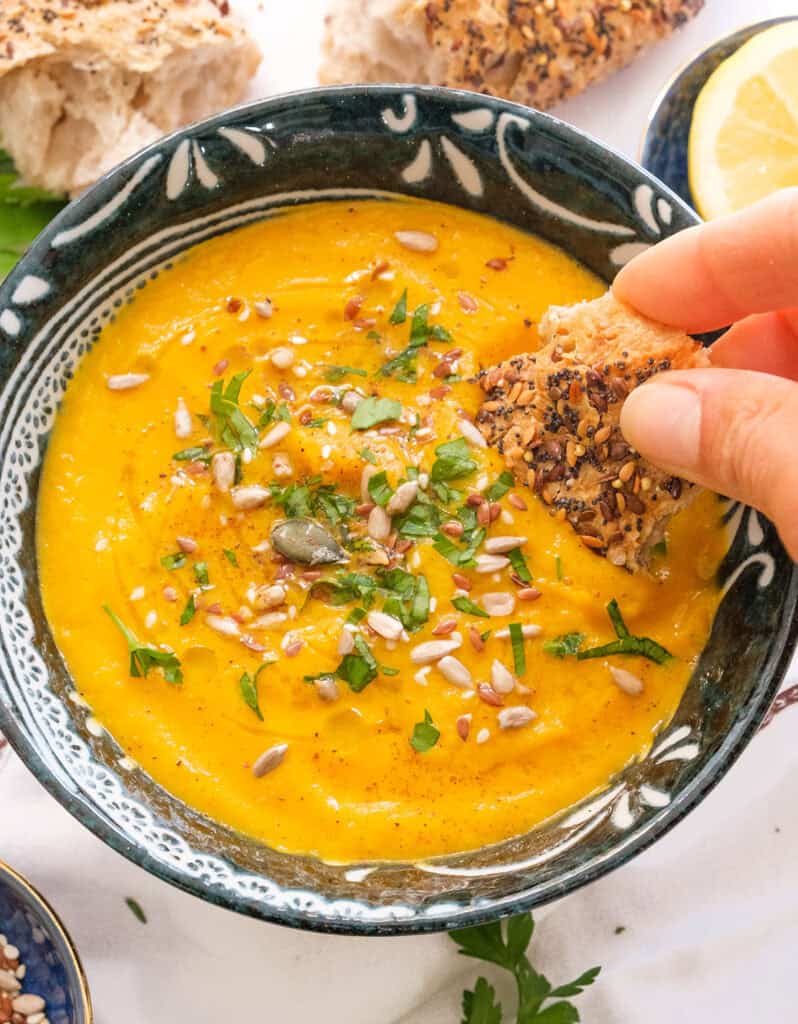 Close-up of a hand dipping some crusty seeded bread into a bowl full of carrot celery soup.