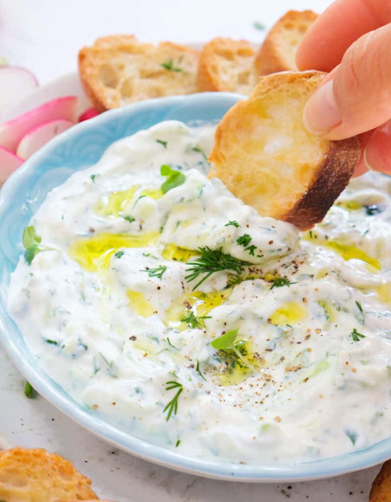 Close-up of crostini dipping into a bowl of vegan tzatziki sauce.