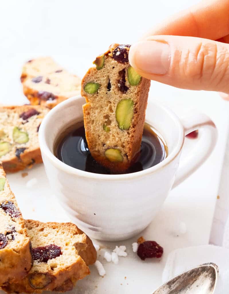 A hand dipping a cranberry pistachio biscotto into a cup of coffee. 